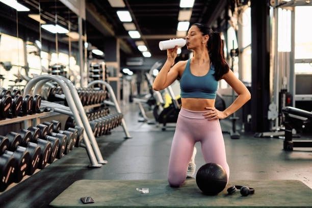Kvinna i gym dricker ur en vit shaker bredvid hantlar